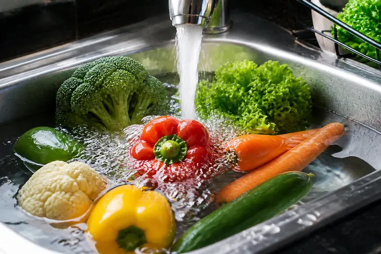 Un gros plan de fruits et légumes lavés sous l'eau courante, illustrant les 3 fruits à éviter pendant la grossesse s'ils ne sont pas propres.